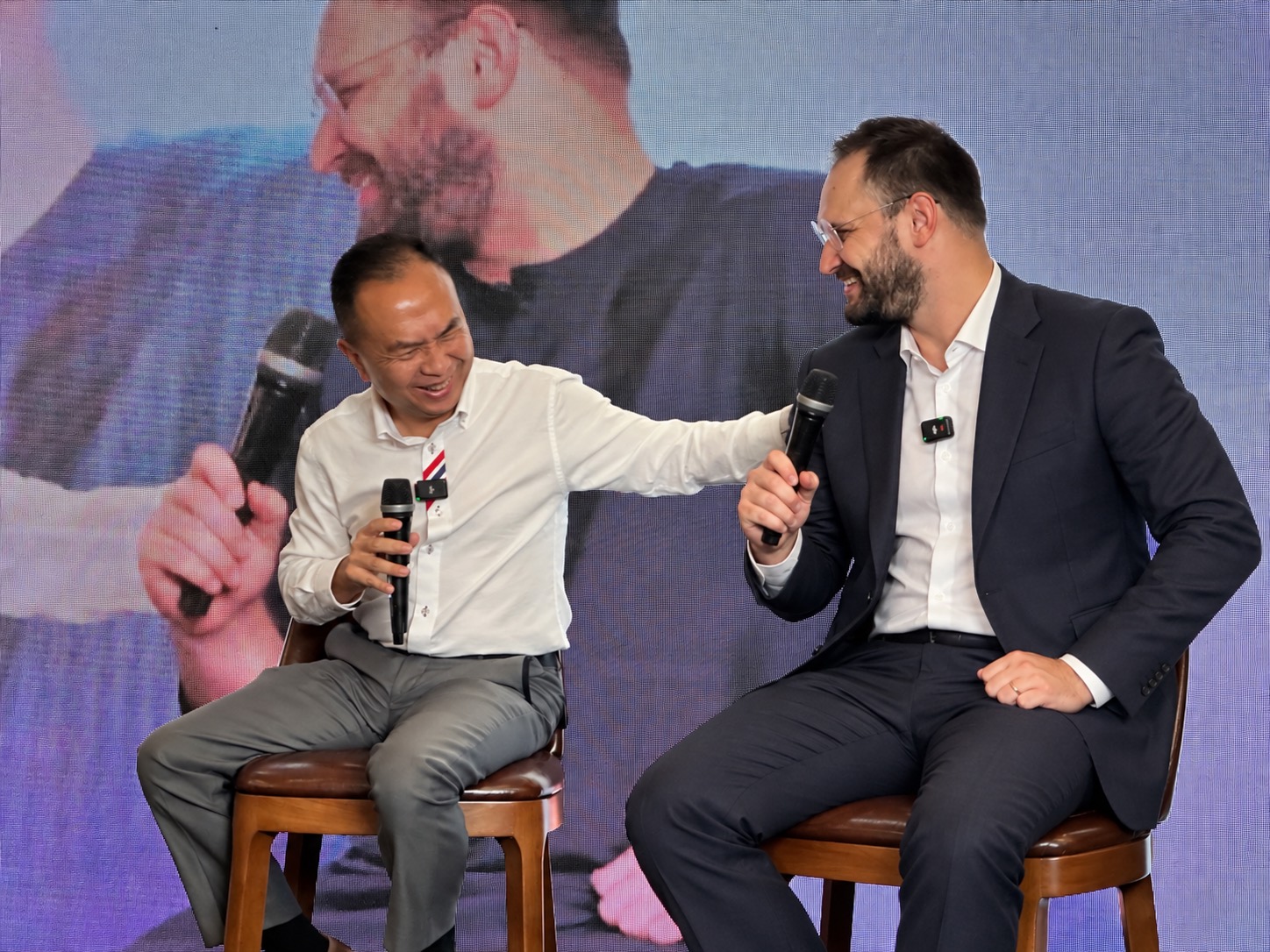 Alex Galt in conversation on stage, both speakers mid-laugh, their faces on the wall behind