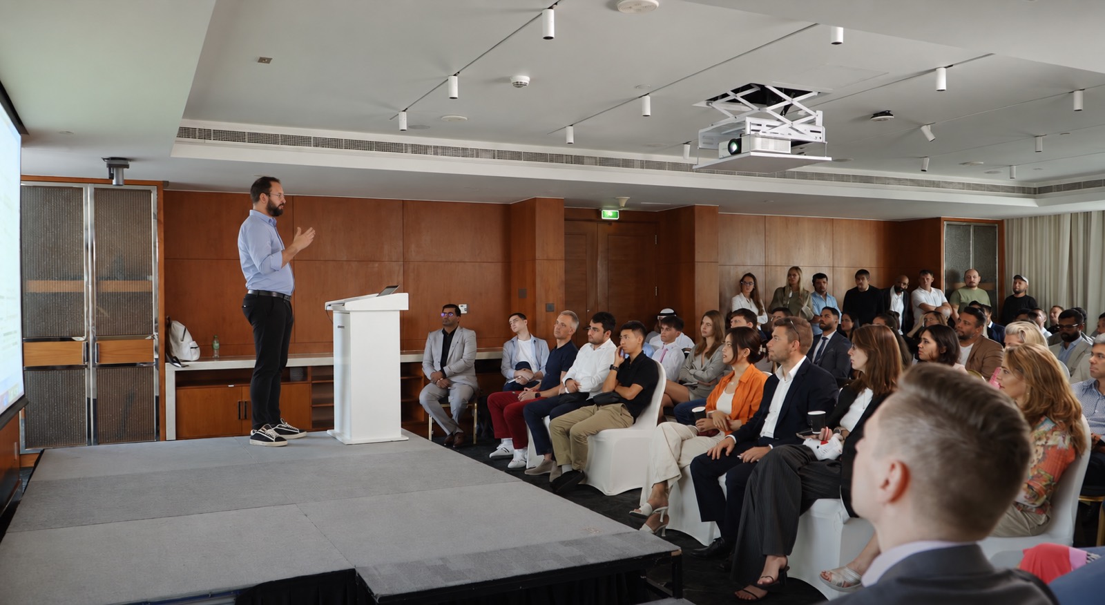 Alex Galt speaking from a lectern to a packed room, the deck on the screen beside him